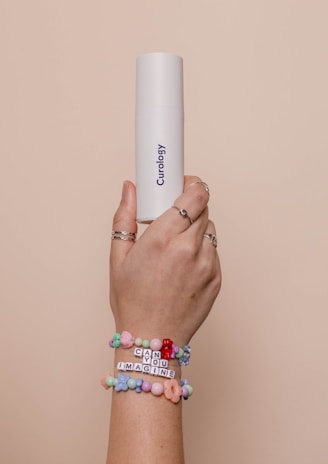 A hand with multiple rings on the fingers is holding a white cylindrical container labeled 'Curology'. The wrist is adorned with colorful beaded bracelets, one of which spells out 'CAN YOU IMAGINE'. The background is a soft beige color, creating a neutral setting.