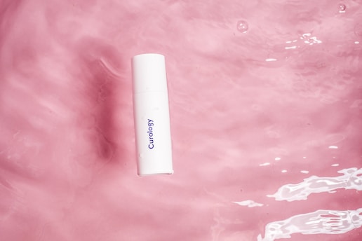 A white bottle with the branding 'Curology' printed in blue lies against a pink watery background. The surface has ripples and reflections, indicating movement and fluidity.