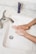 A person is washing their hands under a stream of running water in a white rectangular sink with a silver faucet. A purple tube of lotion or cream is placed on the marble countertop beside the sink.
