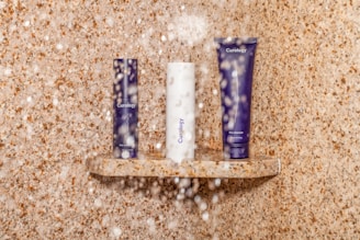 Three skincare products are arranged on a small stone shelf in a shower setting. Water droplets are visible, adding a realistic touch to the scene. The background and shelf feature a beige, speckled stone texture.