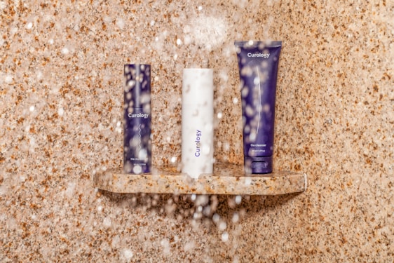 Three skincare products are arranged on a small stone shelf in a shower setting. Water droplets are visible, adding a realistic touch to the scene. The background and shelf feature a beige, speckled stone texture.