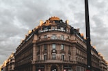 A large, ornate stone building with a round corner and multiple stories is set against a cloudy sky. The building features classic architectural details such as columns and decorative moldings. Signs for 'Royal Air Maroc', 'BERLITZ', and 'BNP Paribas' are prominently displayed. The scene has a dramatic lighting effect with hints of sunlight on the building's edges, creating a contrast with the overcast sky.