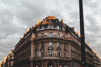 A large, ornate stone building with a round corner and multiple stories is set against a cloudy sky. The building features classic architectural details such as columns and decorative moldings. Signs for 'Royal Air Maroc', 'BERLITZ', and 'BNP Paribas' are prominently displayed. The scene has a dramatic lighting effect with hints of sunlight on the building's edges, creating a contrast with the overcast sky.