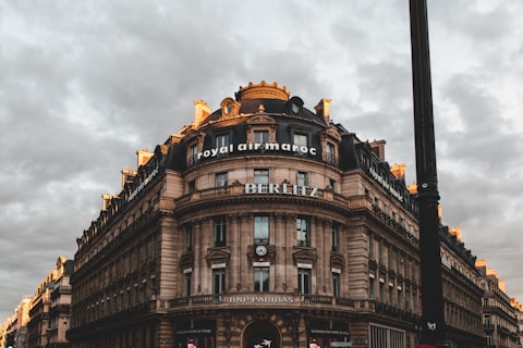 A large, ornate stone building with a round corner and multiple stories is set against a cloudy sky. The building features classic architectural details such as columns and decorative moldings. Signs for 'Royal Air Maroc', 'BERLITZ', and 'BNP Paribas' are prominently displayed. The scene has a dramatic lighting effect with hints of sunlight on the building's edges, creating a contrast with the overcast sky.