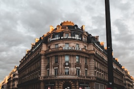 A large, ornate stone building with a round corner and multiple stories is set against a cloudy sky. The building features classic architectural details such as columns and decorative moldings. Signs for 'Royal Air Maroc', 'BERLITZ', and 'BNP Paribas' are prominently displayed. The scene has a dramatic lighting effect with hints of sunlight on the building's edges, creating a contrast with the overcast sky.