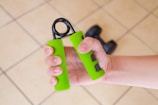 A hand is gripping a green hand exercise tool with a partially visible dumbbell in the background, set against a tiled floor.
