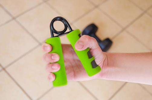 A hand is gripping a green hand exercise tool with a partially visible dumbbell in the background, set against a tiled floor.