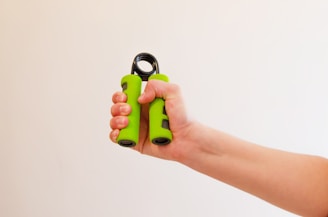 A hand squeezing a green hand grip exerciser, with the arm extended against a plain, light-colored background.
