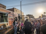 A bustling outdoor market scene with people lining up at food trucks. The sun is setting, casting a warm glow over the crowd. Various food trucks are parked on a brick-paved street with a mix of buildings in the background. Families and friends are gathered, some with children, enjoying the festive atmosphere.