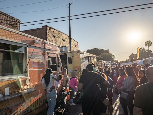 A bustling outdoor market scene with people lining up at food trucks. The sun is setting, casting a warm glow over the crowd. Various food trucks are parked on a brick-paved street with a mix of buildings in the background. Families and friends are gathered, some with children, enjoying the festive atmosphere.