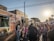 A bustling outdoor market scene with people lining up at food trucks. The sun is setting, casting a warm glow over the crowd. Various food trucks are parked on a brick-paved street with a mix of buildings in the background. Families and friends are gathered, some with children, enjoying the festive atmosphere.