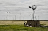 A landscape with flat terrain and a cloudy sky features numerous wind turbines spread across the horizon. In the foreground, there is an old metal windmill atop a small, dilapidated structure surrounded by some green vegetation.