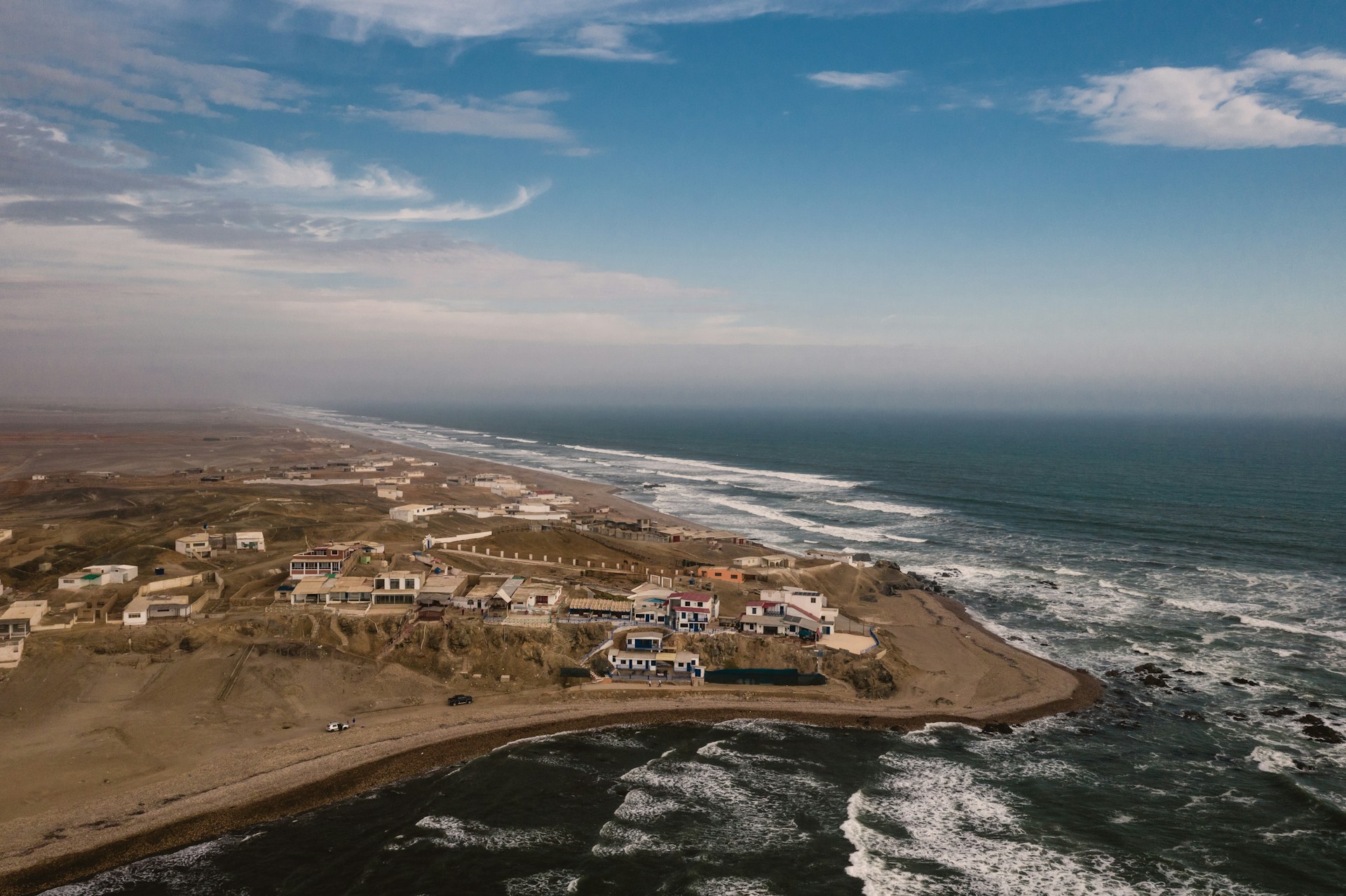 A coastal landscape with several houses perched on a rugged cliff. The ocean waves crash against the rocky shore, and a vast stretch of coastline extends into the distance. The sky is partly cloudy, adding texture to the blue expanse above.