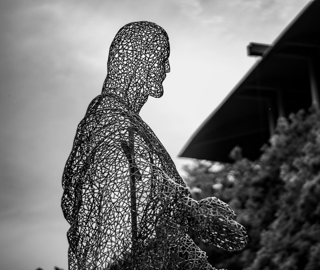 A wire mesh sculpture forms the profile of a person against a cloudy sky backdrop. The structure is intricate, with numerous intersecting lines creating a detailed outline. In the background, there is a building with modern architectural elements and some blurred greenery.