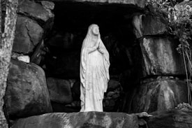 A statue of a woman in a praying position, draped in a long robe, standing within a rocky alcove. The setting appears to be serene and sacred, with large rocks and some foliage seen around the statue.