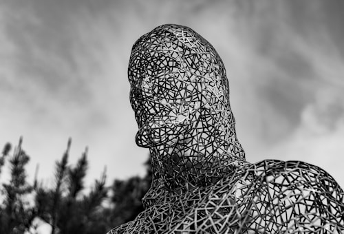 A large metal sculpture of a human head and shoulders, made from an intricate lattice of metal strips, set against a cloudy sky. The open framework allows light to pass through, creating an interplay of shadows and highlights.