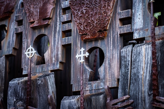 A close-up of an artistic wooden sculpture featuring a detailed carving with two cross-like symbols. The wood appears weathered with a textured surface of various shades of brown and grey. There are intricate cutouts and patterns visible in the design.