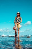 A woman wearing a wide-brimmed natural straw hat, smiling under bright sunlight near the ocean.