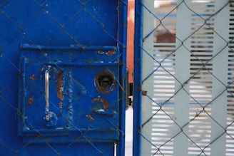 A metal gate with blue paint, showing signs of rust and wear, is partially open. The gate features a silver handle and a keyhole. A chain-link fence is interlaced with the gate, and there's a perforated panel to the right side.
