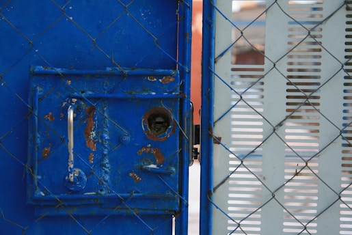 A metal gate with blue paint, showing signs of rust and wear, is partially open. The gate features a silver handle and a keyhole. A chain-link fence is interlaced with the gate, and there's a perforated panel to the right side.