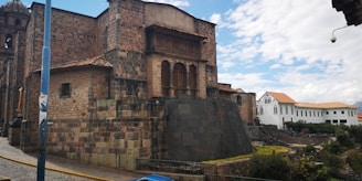 A large, historic stone building with intricate masonry and a distinct architectural design. The structure features a mix of large stone blocks and smaller bricks, with arched windows and a wooden balcony. In the background, there is a newer white building with a tiled roof. The sky is partly cloudy, and some greenery is visible near the base of the buildings.