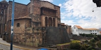 A large, historic stone building with intricate masonry and a distinct architectural design. The structure features a mix of large stone blocks and smaller bricks, with arched windows and a wooden balcony. In the background, there is a newer white building with a tiled roof. The sky is partly cloudy, and some greenery is visible near the base of the buildings.