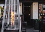 A tall outdoor heater with a visible flame enclosed in a glass cylinder is situated on a patio. The heater is positioned in front of an open doorway leading into a building with white brick walls. The surrounding area includes wooden chairs and tables, indicating an outdoor seating space.