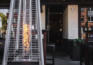 A tall outdoor heater with a visible flame enclosed in a glass cylinder is situated on a patio. The heater is positioned in front of an open doorway leading into a building with white brick walls. The surrounding area includes wooden chairs and tables, indicating an outdoor seating space.