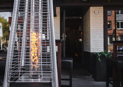 A tall outdoor heater with a visible flame enclosed in a glass cylinder is situated on a patio. The heater is positioned in front of an open doorway leading into a building with white brick walls. The surrounding area includes wooden chairs and tables, indicating an outdoor seating space.