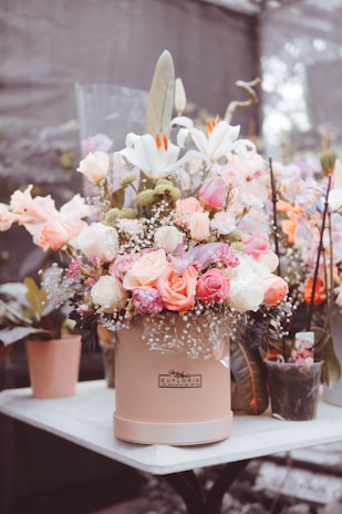 A large bouquet of flowers in a round beige box is placed on a white table. The bouquet contains a variety of flowers, including white lilies, pink and peach roses, and other smaller flowers with green filler greenery. Some additional potted plants and flowers are present in the background.