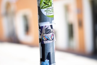 A metal pole is covered with various colorful stickers, including one with a vibrant bikini design and others with graphic and text elements. The background is blurred, hinting at an urban setting.
