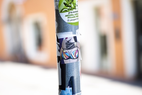 A metal pole is covered with various colorful stickers, including one with a vibrant bikini design and others with graphic and text elements. The background is blurred, hinting at an urban setting.