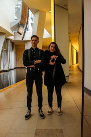 Two individuals are standing in a modern architectural setting with a reflective surface nearby. One person is holding a camera, suggesting an informal or candid moment. The architecture features angled lines and large glass elements, creating an open and airy space.