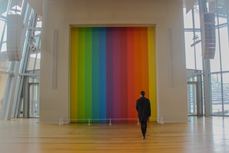 A person in a dark coat stands in a spacious, modern interior with wooden flooring, facing a large wall adorned with vertical stripes of vibrant colors ranging from green to red.