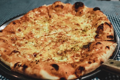 A freshly baked pizza with a golden-brown crust topped with melted cheese and herbs. The pizza is placed on a silver metal tray, and the background features a blue and white checkered tablecloth.