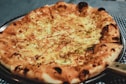 A freshly baked pizza with a golden-brown crust topped with melted cheese and herbs. The pizza is placed on a silver metal tray, and the background features a blue and white checkered tablecloth.