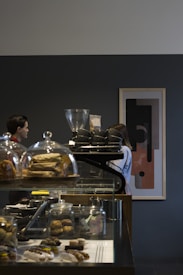 A modern coffee shop interior with a dark wall featuring abstract art. In the foreground, a glass display case contains various pastries such as croissants and scones. Two people are partially visible behind the counter engaged in coffee-making activities, with one using a coffee grinder.