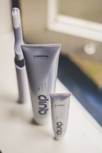 A set of oral hygiene products is displayed on a windowsill. The focus is on a toothbrush and two tubes of toothpaste labeled with time periods, '3 MONTHS' on the larger tube and '2 WEEKS' on the smaller one. The design is minimal with a soft, neutral background and gentle lighting.