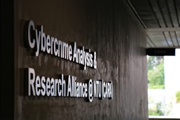 Signage on a wall reads 'Cybercrime Analysis & Research Alliance @ NTU (CARA)' in bold, white letters against a dark, textured surface. A corridor with partial ceiling panels is visible, and there's some greenery in the background.
