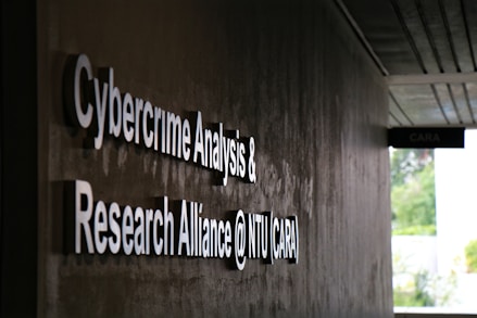 Signage on a wall reads 'Cybercrime Analysis & Research Alliance @ NTU (CARA)' in bold, white letters against a dark, textured surface. A corridor with partial ceiling panels is visible, and there's some greenery in the background.