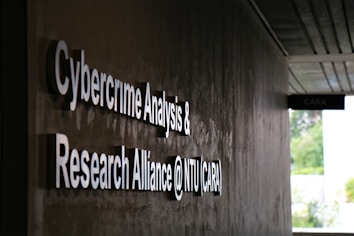 Signage on a wall reads 'Cybercrime Analysis & Research Alliance @ NTU (CARA)' in bold, white letters against a dark, textured surface. A corridor with partial ceiling panels is visible, and there's some greenery in the background.