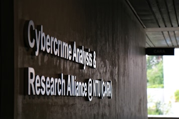 Signage on a wall reads 'Cybercrime Analysis & Research Alliance @ NTU (CARA)' in bold, white letters against a dark, textured surface. A corridor with partial ceiling panels is visible, and there's some greenery in the background.