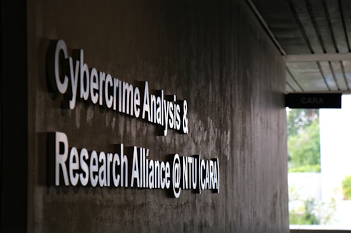 Signage on a wall reads 'Cybercrime Analysis & Research Alliance @ NTU (CARA)' in bold, white letters against a dark, textured surface. A corridor with partial ceiling panels is visible, and there's some greenery in the background.