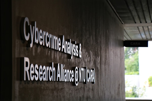 Signage on a wall reads 'Cybercrime Analysis & Research Alliance @ NTU (CARA)' in bold, white letters against a dark, textured surface. A corridor with partial ceiling panels is visible, and there's some greenery in the background.