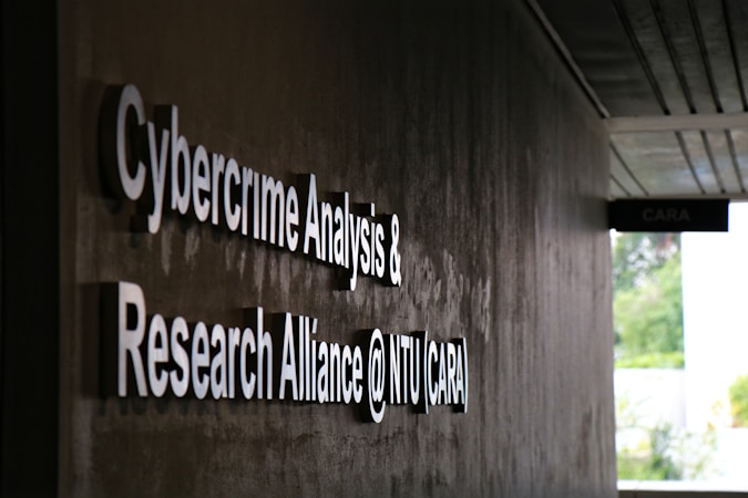 Signage on a wall reads 'Cybercrime Analysis & Research Alliance @ NTU (CARA)' in bold, white letters against a dark, textured surface. A corridor with partial ceiling panels is visible, and there's some greenery in the background.
