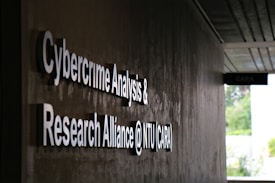 Signage on a wall reads 'Cybercrime Analysis & Research Alliance @ NTU (CARA)' in bold, white letters against a dark, textured surface. A corridor with partial ceiling panels is visible, and there's some greenery in the background.
