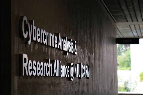 Signage on a wall reads 'Cybercrime Analysis & Research Alliance @ NTU (CARA)' in bold, white letters against a dark, textured surface. A corridor with partial ceiling panels is visible, and there's some greenery in the background.