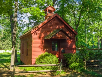 A small, rustic red wooden cabin with a pitched roof is nestled among lush green trees. Sunlight filters through the branches, casting dappled shadows on the cabin. A wooden fence and well-maintained shrubs surround the structure, adding to the serene, natural setting.