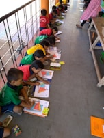 A group of children is sitting on the floor in a line, each engaged in a coloring activity. They are using colorful crayons and focusing on filling in pictures. The scene appears to be in an outdoor or semi-outdoor walkway area, with a railing on one side. The children are wearing brightly colored clothing, which might be uniforms.
