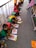 A group of children is sitting on the floor in a line, each engaged in a coloring activity. They are using colorful crayons and focusing on filling in pictures. The scene appears to be in an outdoor or semi-outdoor walkway area, with a railing on one side. The children are wearing brightly colored clothing, which might be uniforms.